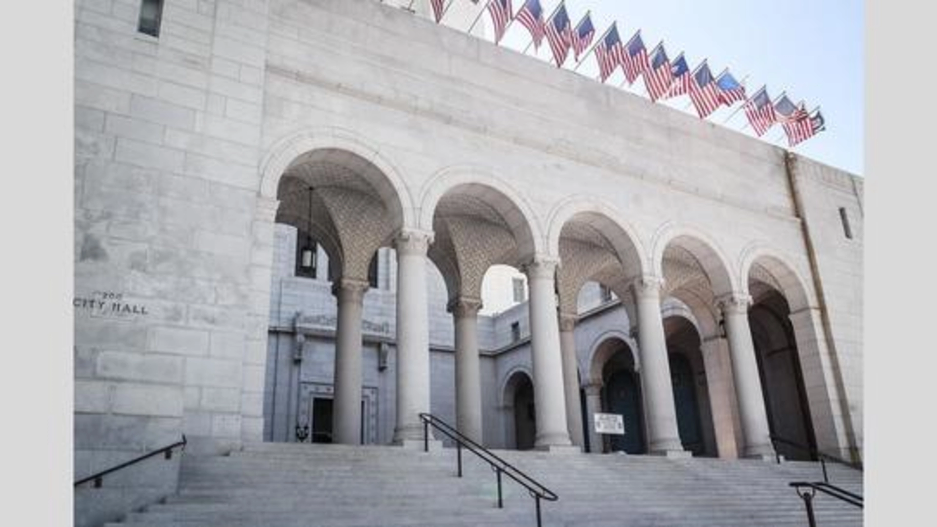 The Los Angeles City Hall forecourt, with stairs leading up, arches and columns and a row of American flags.